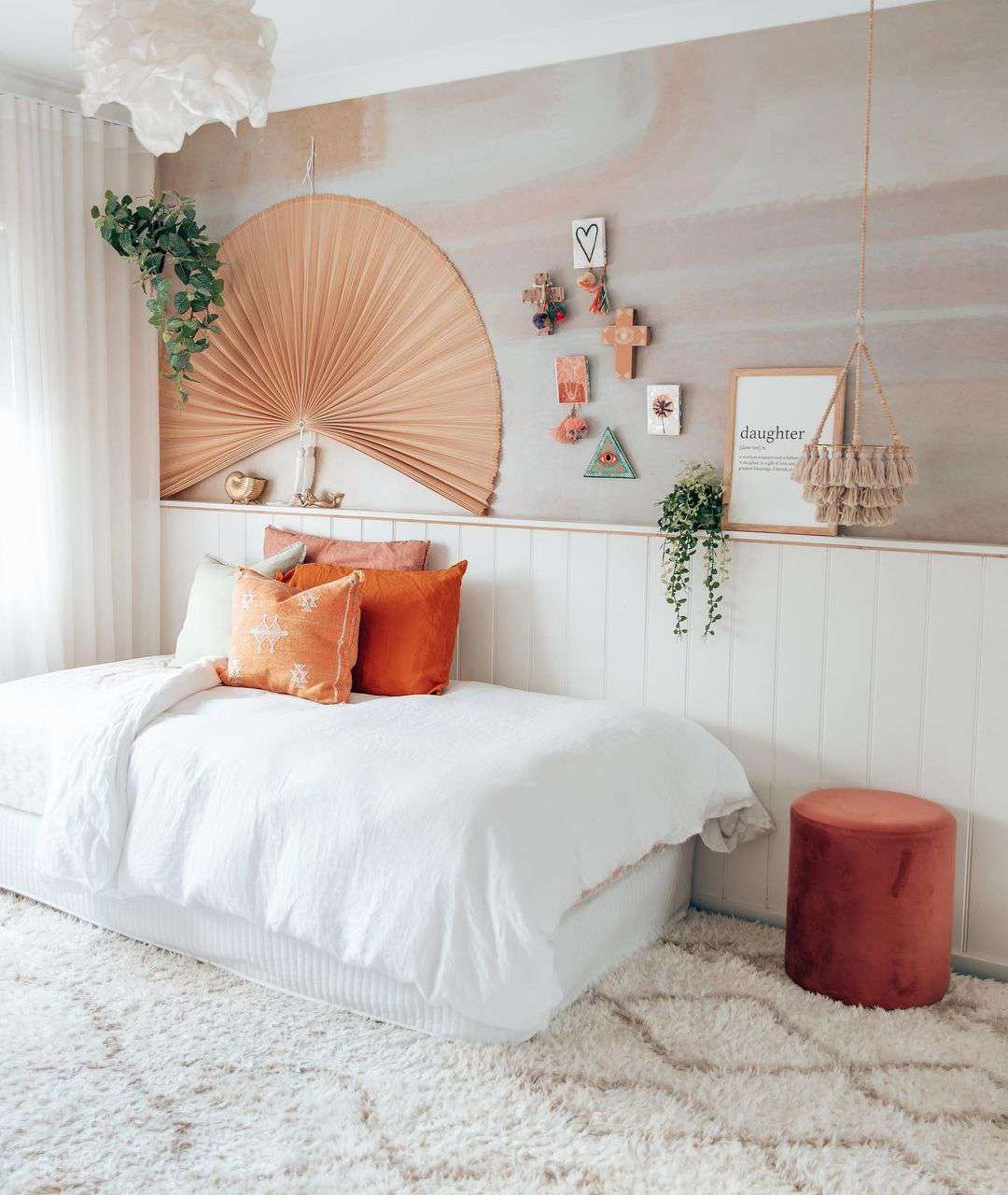 Ένα εφηβικό υπνοδωμάτιο εμπνευσμένο από το boho με τοίχους με ακουαρέλες, έναν μεγάλο χάρτινο ανεμιστήρα, ένα κρεβάτι ημέρας και μια κρεμαστή γλάστρα με φούντες.